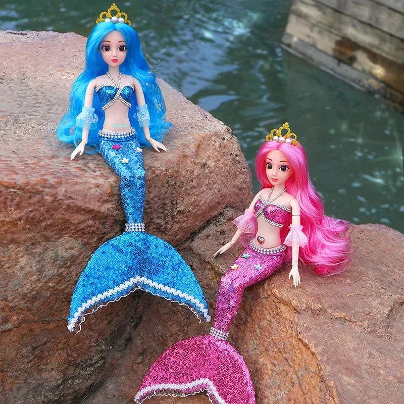Two mermaid dolls with blue and pink tails sitting on a rock by water.
