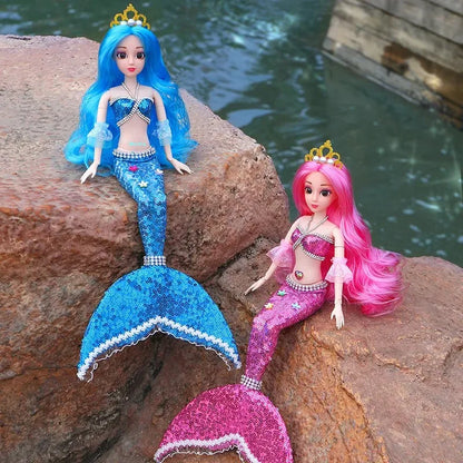 Two mermaid dolls with blue and pink tails sitting on a rock by water.